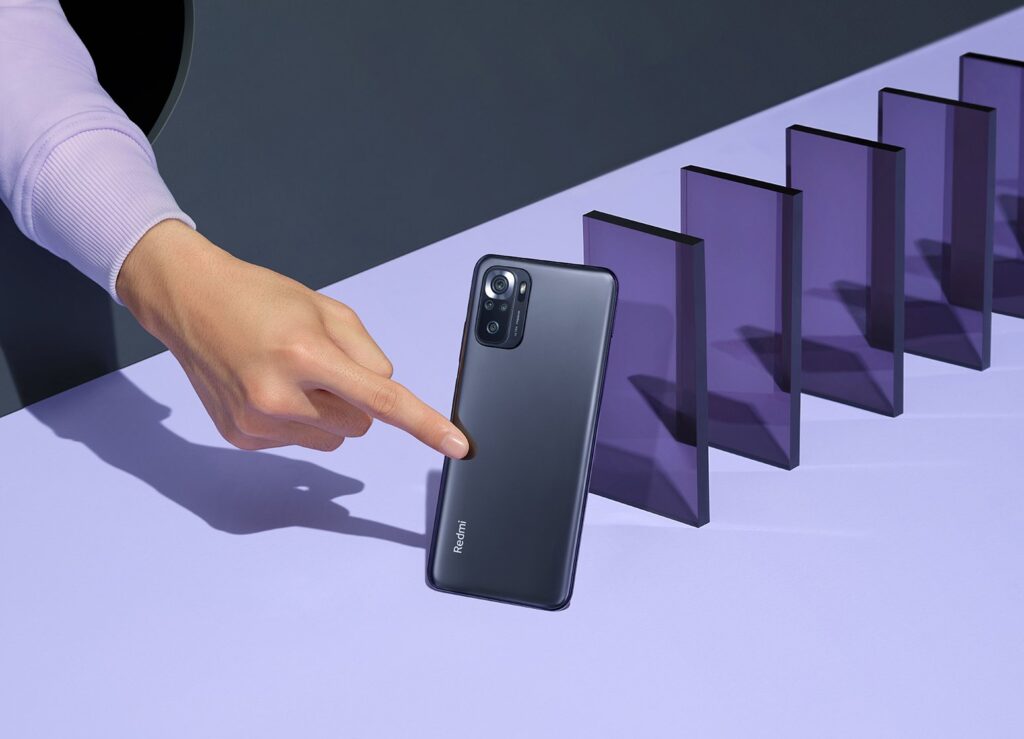 A finger taps the onyx gray Redmi phone like a domino into a series of similar purple translucent shapes, on a lilac background