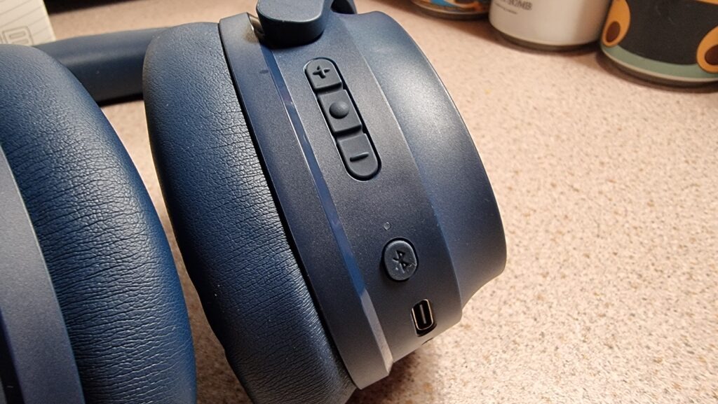 A close-up photograph of the Edifier WH700NB wireless headphones in navy blue; showing the on-board buttons, including the volume up/down buttons, the power button and bluetooth sync button.
