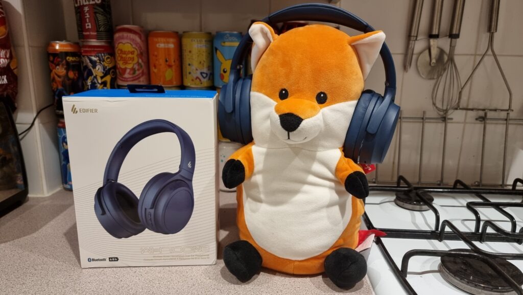 Photograph of the box for the Edifier WH700NB wireless headphones next to a plush fox wearing the headphones on its head. The box and plush fox are sat on a kitchen counter.