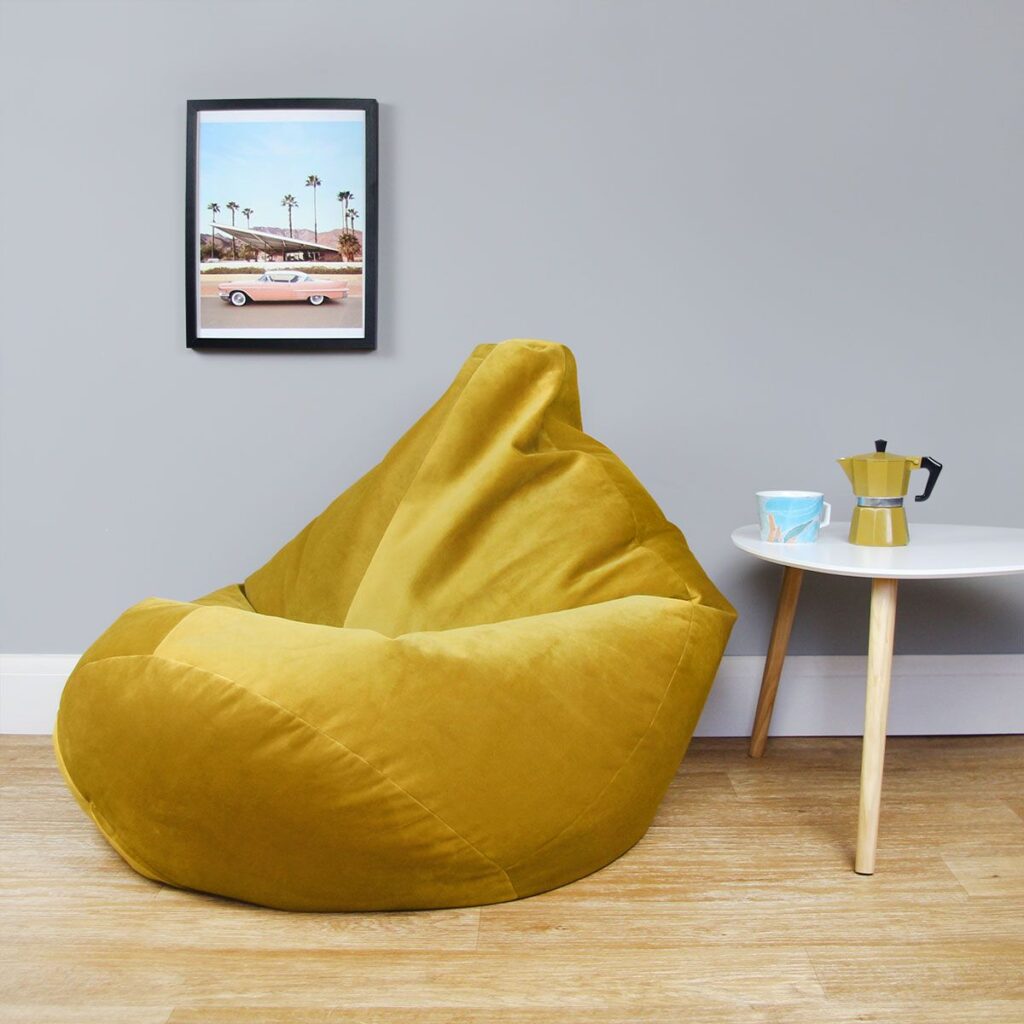 A promotional image of the beanbag in a mustard yellow colour. It is positioned next to a small coffee table adorned with a mug and coffee jug.