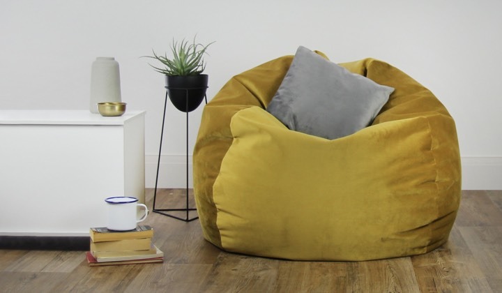 A promotional photograph of the beanbag in mustard yellow in a display room. It has a grey cushion on top of it, and is situated next to a potted plant, a pile of books with a mug on top, and a small cabinet.